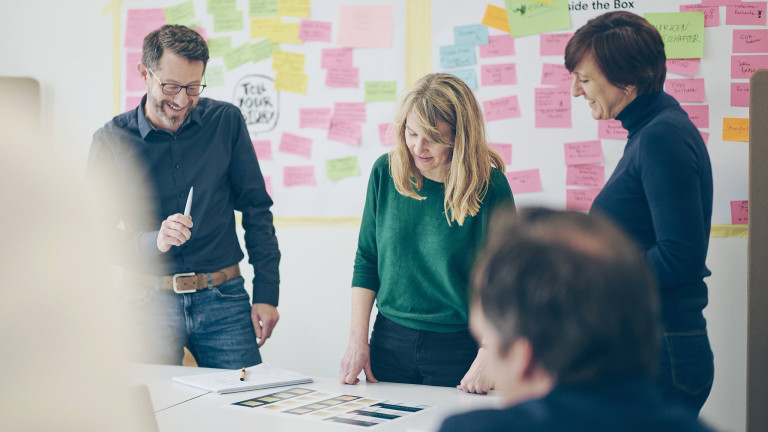 Teammeeting bei DOTS – Die Agentur in Freiburg: Drei Teammitglieder stehen gemeinsam an einem Tisch, umgeben von bunten Post-its an der Wand. Sie diskutieren kreative Konzepte und prüfen Designideen, um innovative Kommunikationslösungen zu entwickeln. Das Bild spiegelt die kollaborative und dynamische Arbeitskultur der Agentur wider.