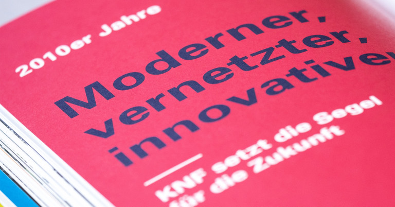 Detailaufnahme einer roten Seite aus der KNF Jubiläumschronik mit der Überschrift: ‚2010er Jahre – Moderner, vernetzter, innovativer. KNF setzt die Segel für die Zukunft.